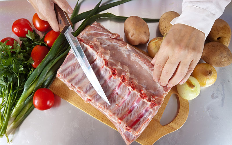 Découpe artisanale d'un carré de côtes de bœuf Halal entouré de légumes frais de saison : tomates, oignons et pommes de terre.